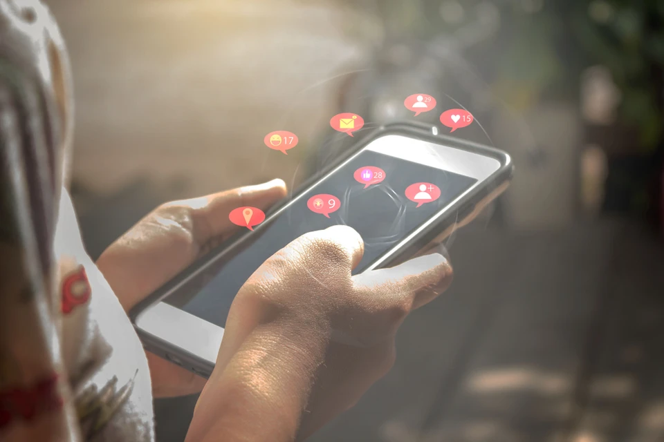 Personne utilisant un smartphone en extérieur, avec des notifications de réseaux sociaux apparaissant au-dessus de l'écran sous forme d'icônes flottantes colorées (likes, messages, abonnés)