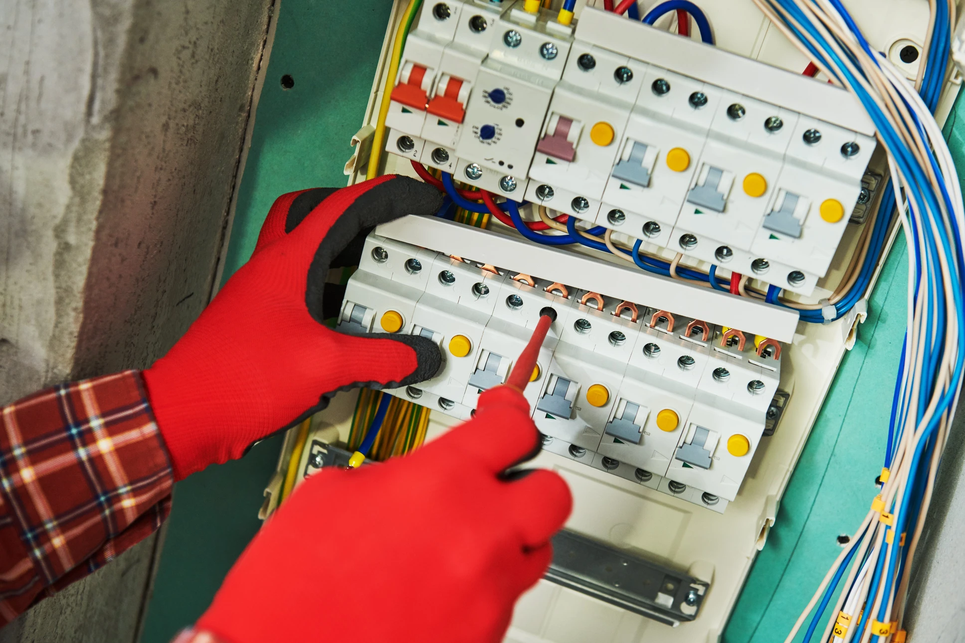 Électricien professionnel souriant travaillant sur un tableau électrique domestique, portant un casque de sécurité blanc et des gants isolants jaunes